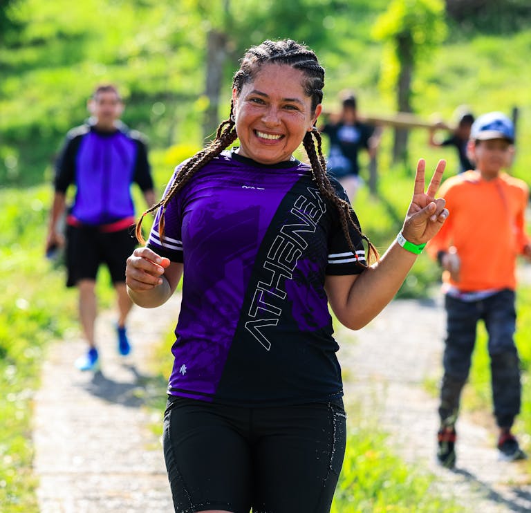 A cheerful female runner flashes a peace sign during an outdoor run with others in a sunny, green setting.