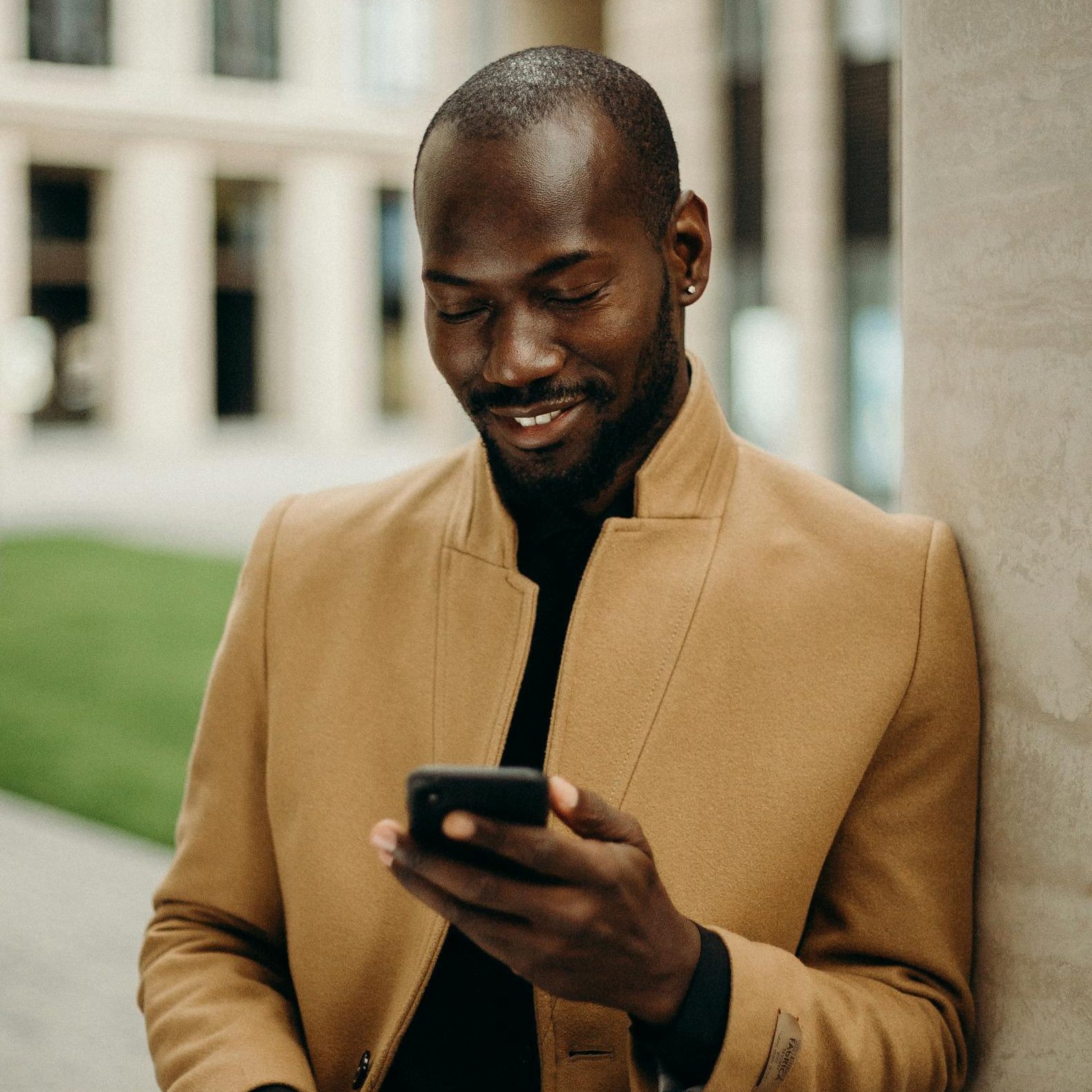 Relaxed man in coat smiles while using smartphone and holding coffee outside.